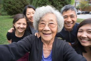 A fit ,senior, Asian Grandmother smiles with her children and mixed-ethnic grandchildren while posing for a selfie in a park garden. Real, three generation family including a senior woman, mature adult son and daughter and teenage grand-daughters. An assisted living facility can be seen in the background. Camera point of view.
